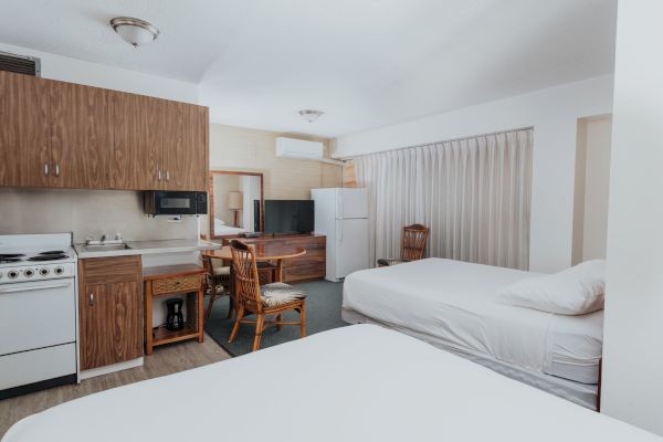 This image shows a room with two beds, a small kitchen area, a mini fridge, a microwave, and a table with two chairs, featuring wooden decor.