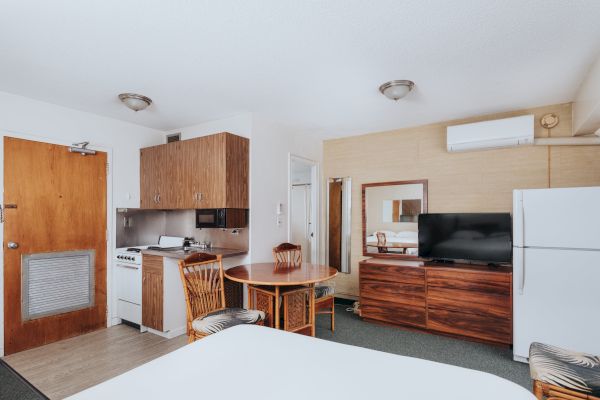 A hotel room with a bed, a kitchenette, a dining table with chairs, a TV, a refrigerator, and a door. The decor is simple and functional.