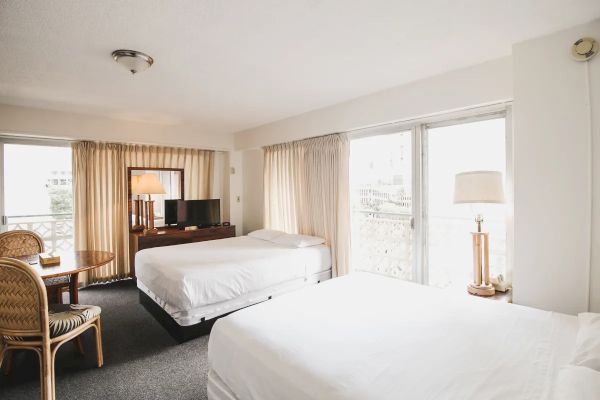 The image shows a bright hotel room with two beds, a TV, a lamp, a small table with chairs, and large windows with curtains.