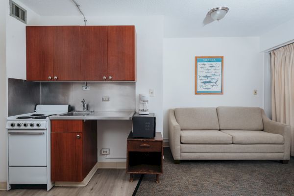 The image depicts a small room with a kitchenette on the left, a beige sofa on the right, and a framed poster of sharks on the wall.