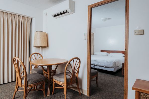 A small dining area with a round table and four chairs is next to an open door leading to a bedroom with a double bed and minimalist decor.
