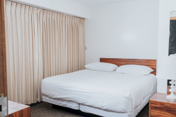 The image shows a minimalist bedroom with a large bed, white linens, beige curtains, and a wooden bedside table with a lamp.
