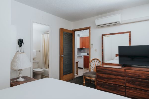 A hotel room with a queen-sized bed, a bathroom, a TV, a wooden dresser, a mirror, an air conditioner, and a kitchenette in the background.