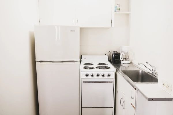 A small kitchen with a refrigerator, stove, sink, coffee maker, and some shelves. The setup is compact and efficient, suitable for limited space.