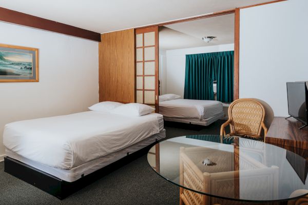 A hotel room with two beds, a glass table, a TV, a wooden chair, blue curtains, and a framed picture on the wall.