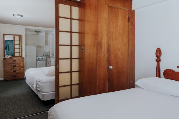 The image shows a bedroom with a wooden sliding door separating it from another room with a dresser, bed, and a small kitchen area in the background.