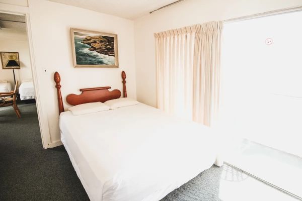 The image shows a bright bedroom with a bed, white linens, a headboard, a seaside painting above, curtains, and a sliding glass door leading outside.