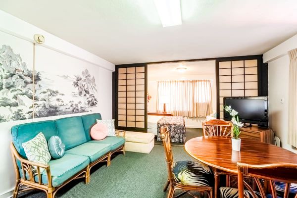 A cozy living area features a teal sofa, decorative pillows, a round wooden table with chairs, and a TV. Sliding doors lead to a bedroom.