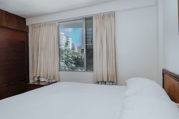 A cozy bedroom with a large bed covered in white linens, beige curtains on a window displaying a view of buildings outside, and wooden furniture.