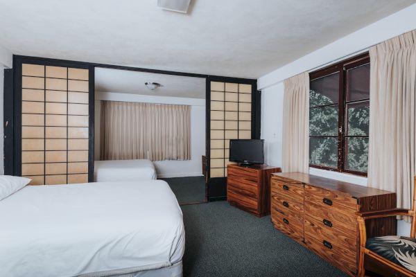 The image shows a bedroom with a bed, a wooden dresser with a TV, and a separate area with another bed, separated by sliding doors.
