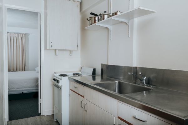 A small kitchen with a sink, stove, and upper shelves holding pots and pans, with a doorway leading to a bedroom with a bed.