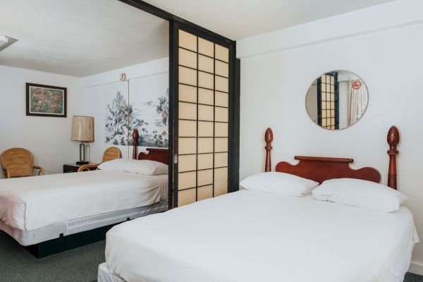A hotel room with two double beds, a circular mirror, framed artwork, wicker chairs, a lamp, and a partition with Japanese-style sliding panels.