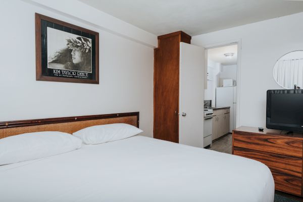 A minimalist bedroom with a bed, a framed picture, a wooden dresser with a TV, and an open door leading to a small kitchen area.