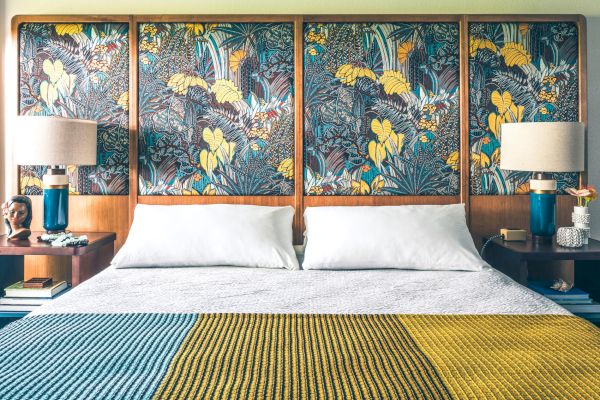 A tidy bedroom with a large bed, vibrant patterned headboard, two bedside tables with lamps, and a colorful knitted blanket on the bed.