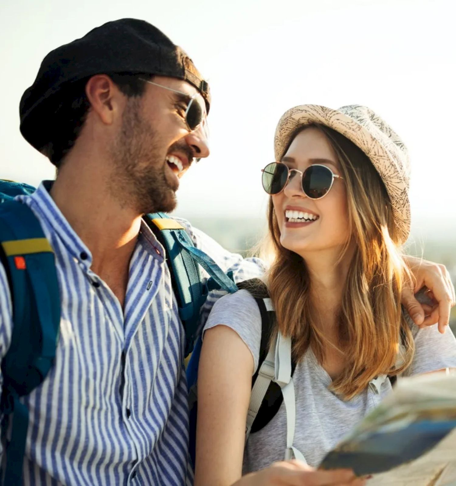 A smiling couple wearing sunglasses and backpacks looks at a map outdoors. They appear to be enjoying a sunny day of exploration.