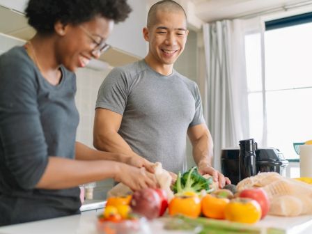 Two people are preparing vegetables together in a bright kitchen while smiling, with various fresh produce on the counter, ending the sentence.