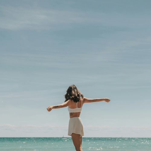 A person stands on the beach with arms outstretched, facing the ocean under a clear blue sky in a serene moment.