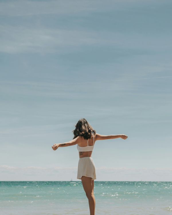 A person stands on the beach with arms outstretched, facing the ocean under a clear blue sky in a serene moment.