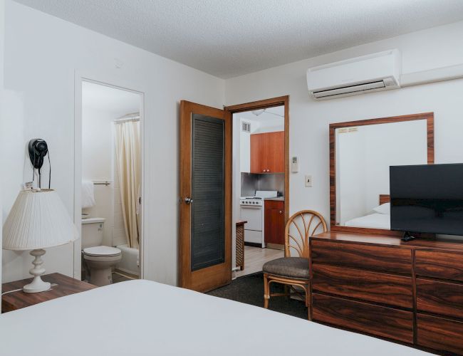 The image shows a tidy hotel room with a bed, dresser, TV, lamp, bathroom, and kitchenette, creating a compact and functional living space.